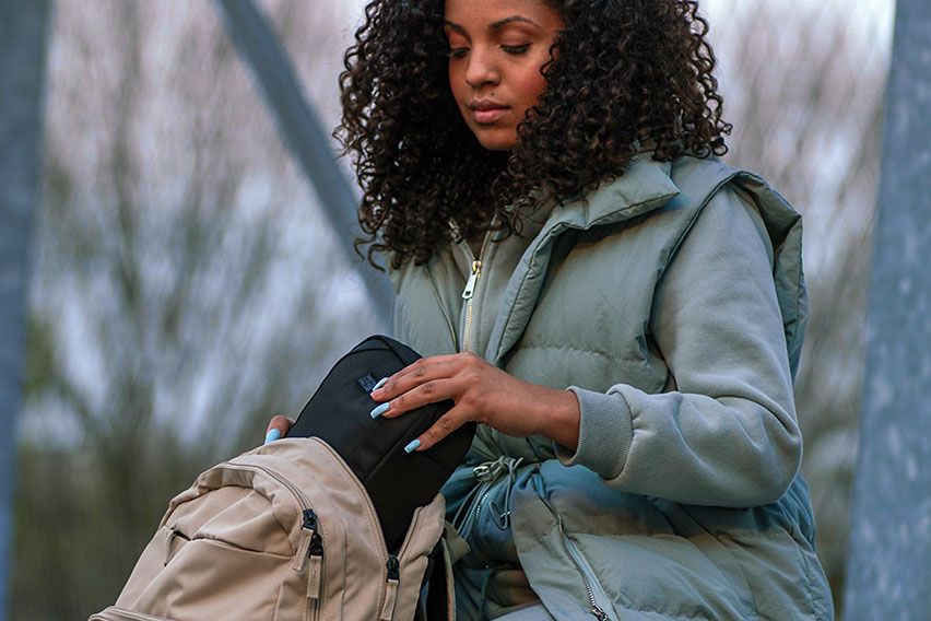 A woman sits in a forest with a blue jacket, placing an item inside a Case Logic backpack for dancers.