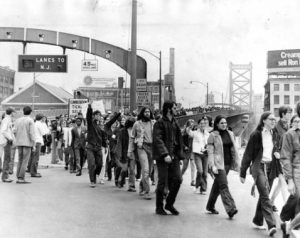 A image of protesters marching from left to right under a bridge arch which holds signal lights for lanes of traffic as well as the speed limit. The bridge extends into the distance to the right of the frame, fading in the atmosphere. Protesters can be seen occupying the walkway as far as can be seen. The suspension towers of the bridge are visible, but very distant.