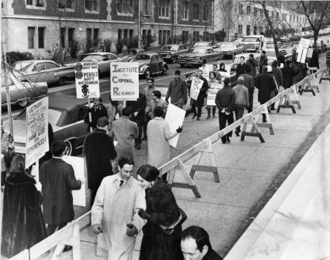 A couple walks arm in arm toward the photographer along the sidewalk at the bottom of the frame. A line of barricades (of wooden saw-horses) stands behind them as marching protesters hold signs in the street near parked cars. Their signs read, 