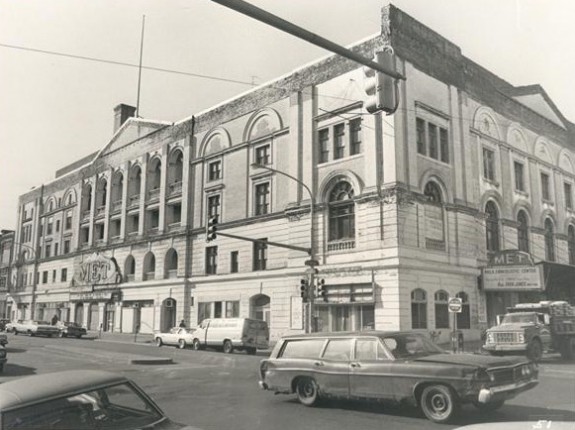 black and white photograph of the metropolitan opera house, taken from the adjacent corner of broad and poplar streets. The building spans almost a block. a car passes down broad street in the foreground.