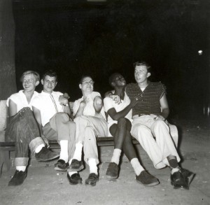 A black and white photograph of a group of five men sitting on a park bench. The background is dark, obscuring any detail. The five people on the bench show various cheerful expressions and are posing for the camera in an exaggerated manner. 