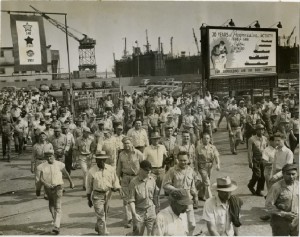 A large crowd of working men in front of the Sun Shipbuilding and Dry Dock Company yards, with a billboard celebrating sixty years in the background