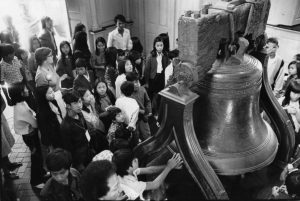 A group of Vietnamese children are gathered around the Liberty Bell while a woman speaks to them. One reaches up to touch the bell. They all have white tour tags hung on lanyards around their necks.