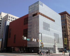 Pictures a large glass and terra-cotta building. In front lies a sculpture and sits an intersection where the sign for 5th Street is visible.