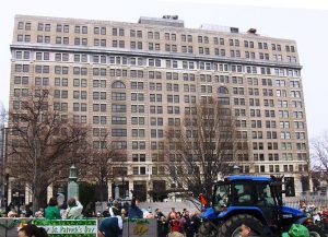 a color photograph of the dupont building in Wilmington with a crowd of people standing in front. A banner in the foreground reads "St. Patricks Day"