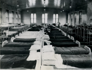 A black and white photograph of a hospital dormitory showing long rows of tightly packed beds, some with patients on them.