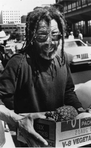 a black and white photograph of John Africa carrying a cardboard box with a pineapple in it.